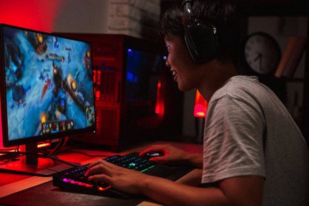 快乐的亚洲青少年玩家的肖像玩电子游戏的电脑在黑暗的房间戴着耳机, 并使用背光彩色键盘