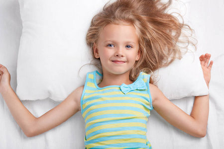 儿童, 床上用品和觉醒的概念。高兴的小欧洲女孩与迷人的温柔的微笑, 长的轻的头发, 穿着 pyjamass, 躺在白色的枕头和床