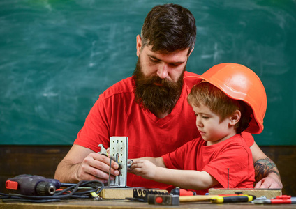 父亲用胡须和小儿子在课堂教学中使用工具, 黑板上的背景。男孩, 孩子在防护头盔做手工, 修理, 做工艺品与爸爸。男士工作理念