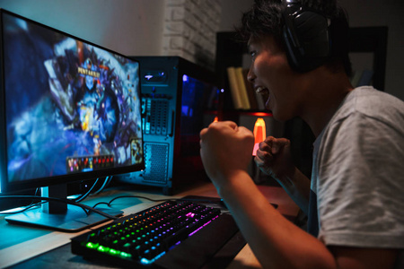 兴奋的亚洲青少年玩家的肖像玩视频游戏在黑暗的房间里戴着耳机和使用背光彩色键盘的计算机上