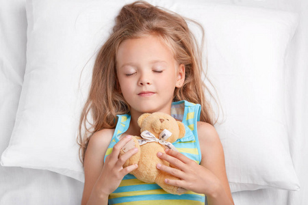 照片的迷人的女孩子晚上有愉快的梦想, 举行泰迪熊, 穿着 pyjamass, 睡在卧室舒适的床上。小孩子带着最喜欢的玩具休息。童