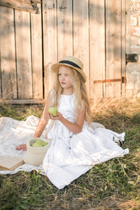 可爱的金发女孩在白色礼服和草帽与苹果在乡下在夏天