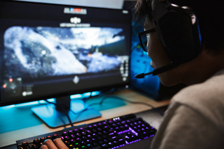 亚洲玩家男孩的照片特写在黑暗的房间里电脑上玩电子游戏, 戴着麦克风的耳机, 使用背光彩色键盘