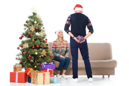 女人看着男人隐藏的心形礼品和站在圣诞树附近, 在白色隔离