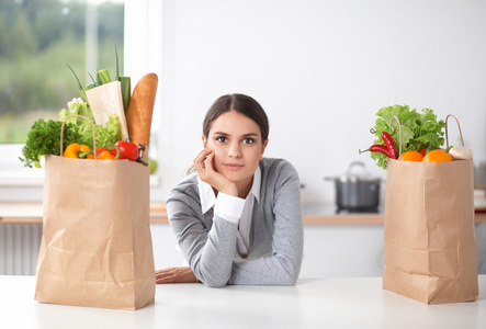 年轻女子捧着与蔬菜站在厨房里的食品杂货店购物袋