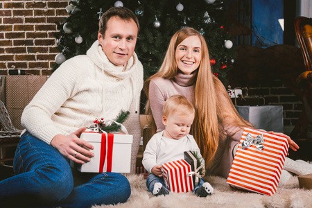家庭庆祝圣诞晚会。家庭三白种人坐在针叶树圣诞树下的地毯羊毛地板上。孩子拿着礼物盒子, 父母年轻时髦穿戴