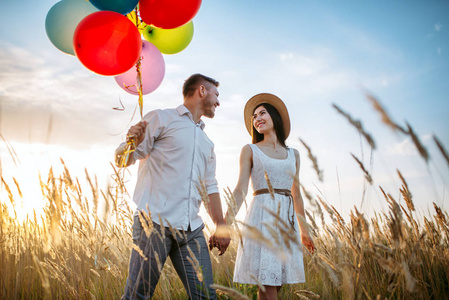 快乐的丈夫和妻子带着一群气球走在麦田里。美丽的夫妇休闲在夏天草甸