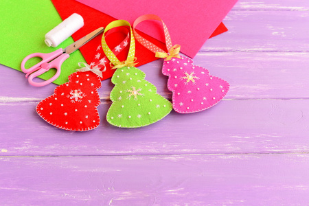 绿色，红色和粉色圣诞树工艺品 剪刀 白线 针，感觉床单上淡紫色的木制背景设置。明亮的圣诞玩具缝毡和装饰珠和雪花