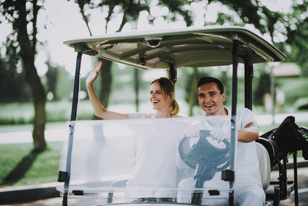 愉快的年轻夫妇在白色购物车在高尔夫球场上。年轻的家庭。高尔夫俱乐部。夏天的运动。在野外的车辆。户外乐趣在夏天。司机与汽车。健康的