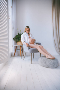 怀孕的 mothercare 生活方式的概念, 鲍勃发型金发女郎的健康护理。白色的灯光内饰, 母亲坐在灰色的扶手椅上, 房间里带
