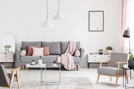 在白色客厅内的扶手椅和桌子, 在灰色沙发上贴上海报和粉红色毯子。真实照片
