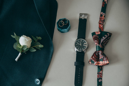 新郎的配件 手表, 绿色夹克, 纽扣, 蝴蝶和戒指在有色米色垫子上