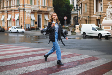 年轻美丽的微笑的女人在皮革夹克和牛仔裤高兴地看着相机捧着杯咖啡走在舒适的城市街道上行走