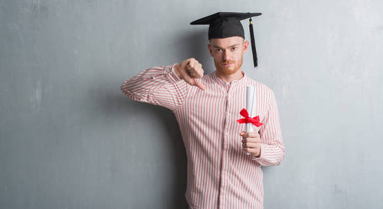 年轻的红发男子在灰色的格格墙上戴着带着愤怒的脸的毕业帽, 负面的迹象显示不喜欢大拇指, 拒绝的概念