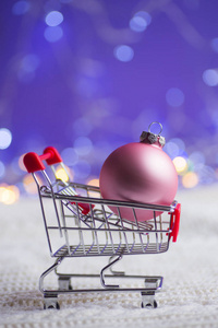 粉红圣诞球在白色针织面料的小超市手推车与花环灯在紫色的波克背景。有空空白的新年横幅。圣诞购物的概念