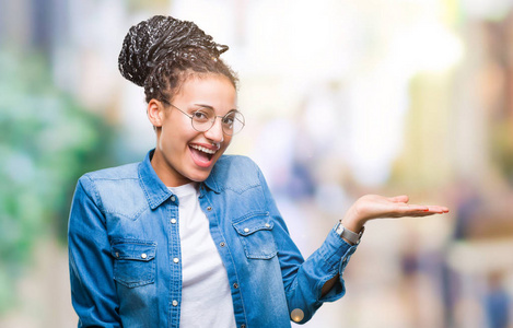 年轻的编织头发非洲裔美国女孩戴眼镜在孤立的背景微笑欢快的呈现和手掌看着镜头
