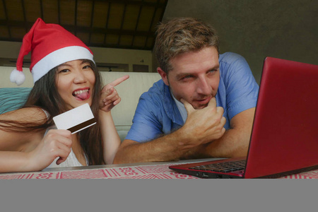 年轻快乐和兴奋的混合族夫妇与亚洲中国妇女在圣诞帽子和白色丈夫网上购物圣诞礼物与信用卡和笔记本电脑在家里的沙发