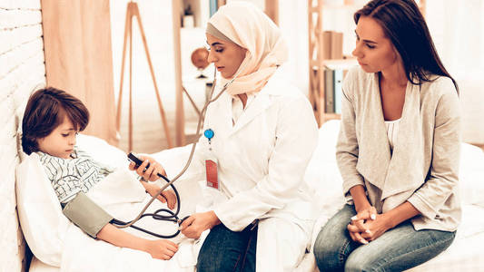 阿拉伯女医生检查血压男孩。儿科医生的孩子。医院理念。健康的理念。儿童病人访问医生。医生检查血压。躺在床上的生病男孩