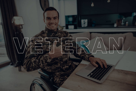 退伍军人。坐在轮椅上的残疾人。坐在桌旁的人。男子使用的笔记本电脑。人是战士。身穿军服的士兵显示好的标志的人。男人的微笑。位于客厅