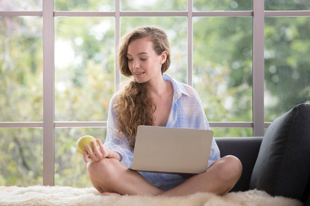 健康的年轻妇女坐在沙发上使用笔记本电脑和持有一个苹果看起来轻松和舒适