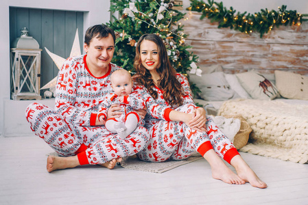 快乐的父母与他们的小女儿在节日的衣服与印刷的鞋和雪花在舒适的房间里, 圣诞树, 礼物和圣诞灯。新年和圣诞节假期概念