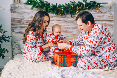 快乐的父母与他们的小女儿与印刷的鞋和雪花打开圣诞礼物在舒适的房间里与圣诞树和圣诞灯