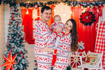 新年和圣诞节假期的概念。父母带着小女儿穿着节日服装, 穿着印刷的子和雪花, 在装饰着圣诞树的房间里, 背景是花圈