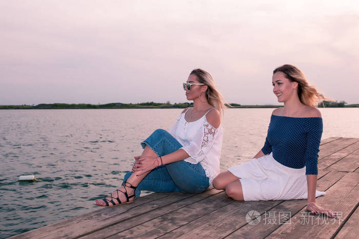 照片 夏天的时间.两个年轻的女性朋友坐在河边.在夏日的傍晚里玩.jpg