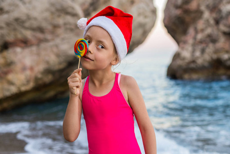 圣诞节背景 可爱的孩子在圣诞老人帽子庆祝新年和圣诞节在海滩上, 自由空间。热带国家的寒假。戴着圣诞帽子的女孩, 戴着棒棒糖