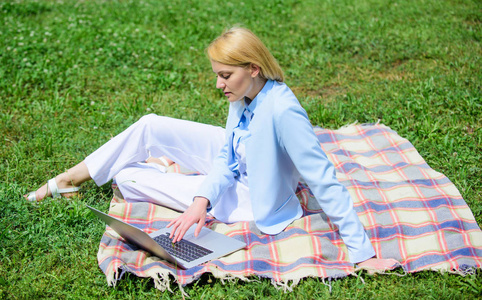 拿着笔记本电脑或笔记本电脑的女人坐在地毯上的绿草草地上。开始自由职业的步骤。商务女士自由职业者在户外工作。在线经营理念。商务野餐