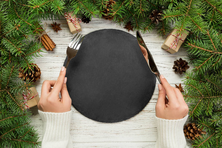 顶视图女孩手里拿着叉子和刀, 准备吃饭。空黑色石板在木圣诞节背景。节日晚餐菜的概念与新年装饰