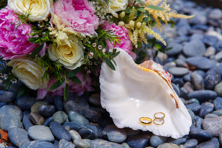 婚礼配饰。一个白色的贝壳与婚礼和订婚戒指躺在海鹅卵石上。背景中新娘的婚礼花束