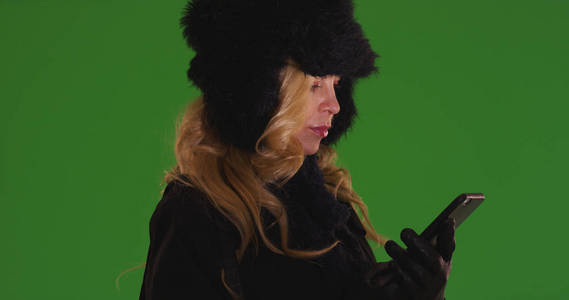 金发碧眼的白色女性在冬季服装使用移动设备在绿色屏幕上