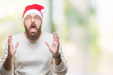 年轻的高加索人戴着圣诞帽, 戴着眼镜在孤立的背景疯狂和疯狂的喊话和大喊大叫, 积极的表情和手臂抬起来。挫折的概念