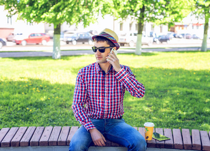 坐在旁边咖啡或茶，通电话，在夏天戴着帽子在树林的户外休息商人谈智能手机在眼镜和一件衬衫的男子