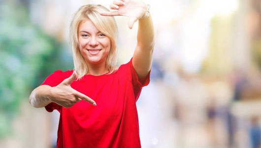 年轻漂亮的金发女人穿着红色 t恤在孤立的背景下微笑着制作框架与手和手指与快乐的脸。创意与摄影理念