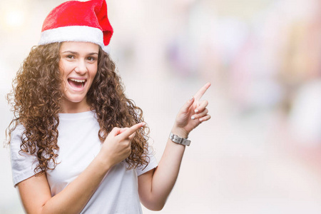 年轻的黑发女孩戴着圣诞帽在孤立的背景微笑着, 看着相机指向与两个手和手指的一侧