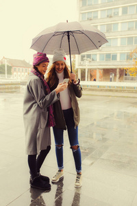 两个年轻快乐的女孩带着伞, 在城里用手机