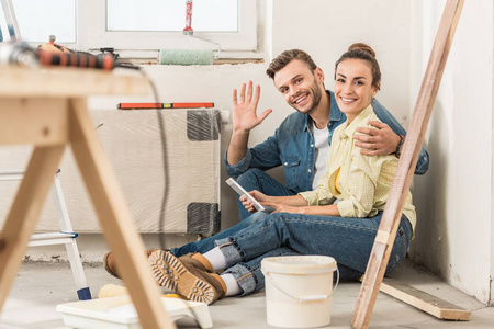 幸福的年轻夫妇使用智能手机和微笑着对着相机, 而坐在地板上在新房子