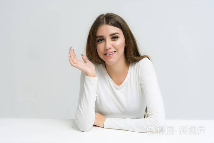 工作室的肖像一个微笑的快乐美丽的黑发女孩在白色背景说话和坐在桌子