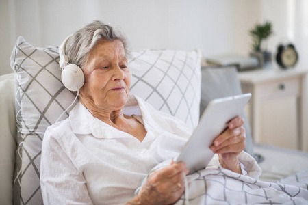 生病的老年妇女, 耳机和平板电脑躺在家里或医院的床上