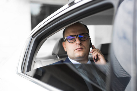 年轻的成功商人坐在一辆精英汽车的后座上打电话, 参加会谈和商务会议