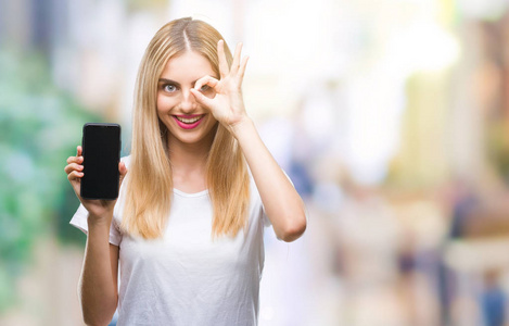 年轻美丽的金发碧眼的女人展示智能手机在孤立的背景与快乐的脸微笑做确定的标志与眼睛看着通过手指