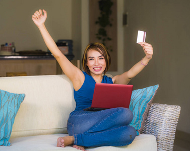 年轻美丽快乐的女孩在家里的客厅拿着信用卡的生活方式肖像使用笔记本电脑银行和网上购物微笑轻松在电子商务和互联网商务