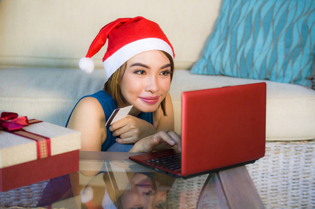 年轻的快乐和美丽的女孩放松在家里的沙发上圣诞老人帽子使用笔记本电脑支付圣诞礼物与信用卡微笑在网上购物和互联网商务