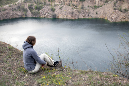 女性坐在大石读物的蓝色的湖面。对银行的岩石山麓。拥抱她的双腿