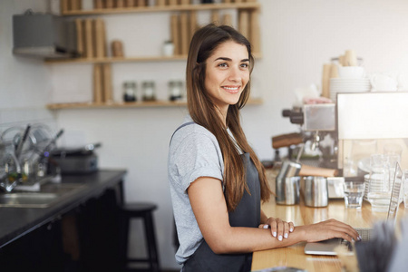 年轻女人咖啡师在她工作场所看快乐微笑在笔记本电脑上工作