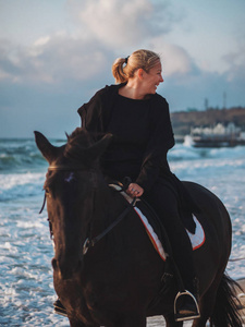 shof 的女人坐在那匹黑马在海上海滩。美丽的女人，穿着黑色的衣服，享受日出早晨接近海洋水