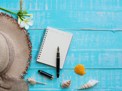 海滩配件包括鲜花, 帽子海滩, 贝壳, 笔记簿和钢笔在明亮的蓝色粉彩木背景为暑假和假日概念