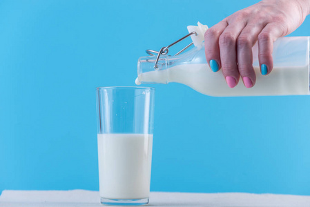 一个女人的手把新鲜的牛奶从玻璃瓶倒入蓝色背景的玻璃杯中。多彩简约。健康乳制品与钙的概念